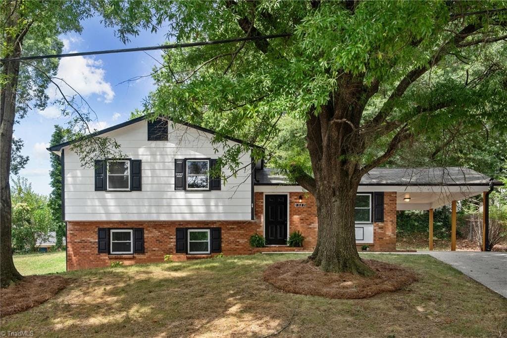 Front exterior view showcasing a spacious, well-maintained yard and a beautiful mature tree providing ample shade. The house has a charming traditional style with a welcoming entrance.